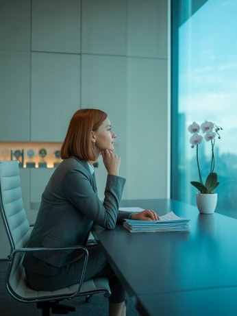 אישה חושבת מול חלון במשרד – דימוי לאישה מצליחה שמבקשת יותר איזון פנימי דרך אימון אישי לחיים